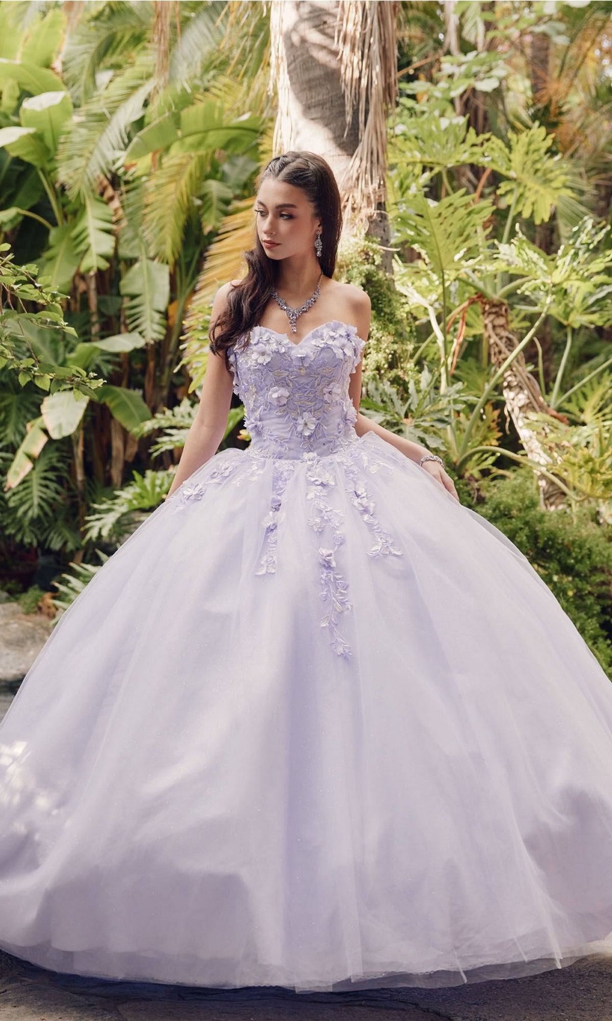 A woman stands outdoors in front of lush green plants and palm trees, wearing the Juliet 1435 Strapless Ball Gown Quinceañera Dress in lavender with floral appliqués.